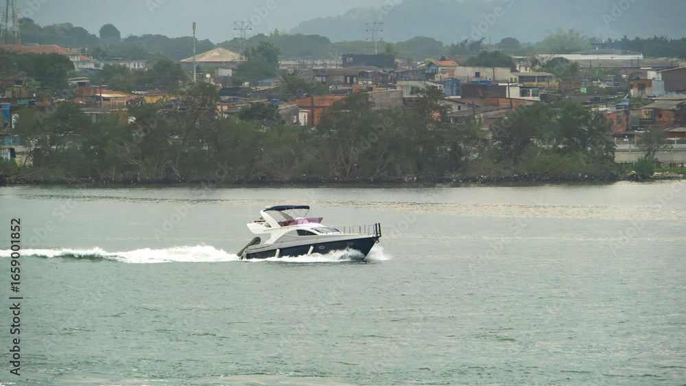 Obraz premium Luxury motor yacht speeds across the water near a coastal town on a bright day, leaving a white wake