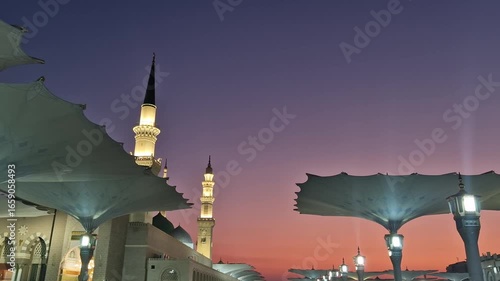 Pan left of giant umbrellas, green dome and minarets of Nabawi mosque during early morning sunrise in Medina, Kingdom of Saudi Arabia.