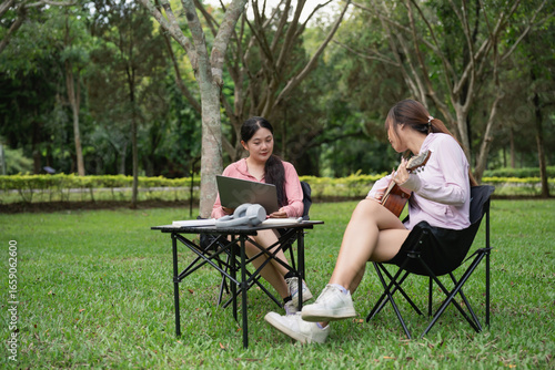Two Young Women Enjoy Outdoor Leisure Time in a Park, One Working on Laptop While the Other Plays Guitar in a Serene Natural Setting