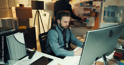 Man, police and research on computer in office for criminal records, case report or problem solving. Night, detectives and tech in precinct for crime investigation, evidence analysis or trace suspect