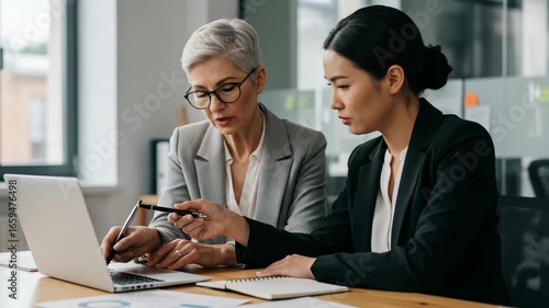 Two professional women, one senior and one younger, collaborating on a laptop during a business meeting, focusing on strategy and analysis