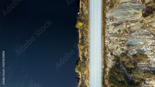 Top down view of a beautiful road in Norway (Mykland) next to a dark blue lake. Rocks and a few trees on the right and left side of it.