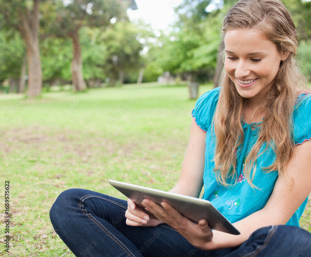 Fototapeta premium Woman sitting cross-legged on grass in public park holding tablet and smiling, copy space