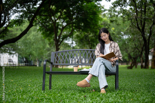 Young asian glasses woman holding pencil writing in notebook as sitting on bench in park or garden