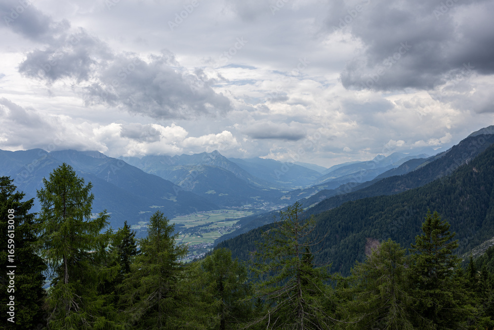 Obraz premium Wide view of alpine valley with mountains, forest slopes, and cloudy dramatic sky above