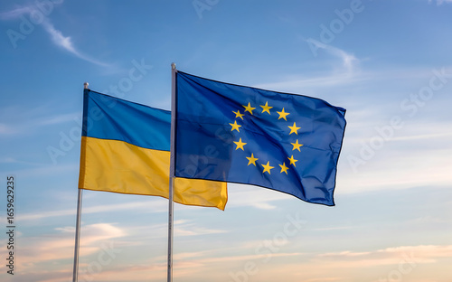 A photograph of two flags waving gently in a clear blue sky, the flags representing Ukraine and the European Union. The Ukrainian flag features horizontal bands of blue and yellow, while the European 