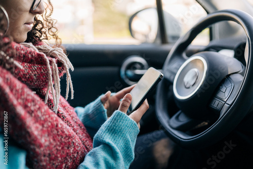A woman with curly hair and glasses is seated inside a car, actively using her smartphone. She wears a warm red scarf and a blue sweater, focused on the device.