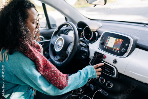 A young woman with curly hair and glasses sits in the driver's seat of a car. She is using her smartphone, which is mounted on the dashboard. The car's interior is visible.