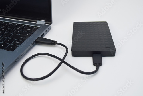 External hard drive connected to laptop with USB cable on white desk, concept of portable storage, data transfer, and backup technology.