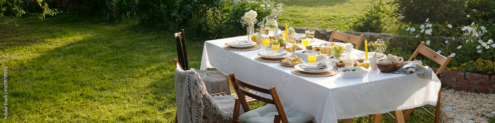 Fototapeta premium Outdoor dining table set for meal with plates, glasses, bread, salads, juice, flowers arranged on tablecloth, wooden chairs surrounding table in garden with green grass