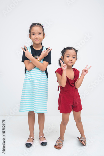 A pair of cheerful young asian girls posing playfully together, standing next to each other with wide smiles while raising both hands in peace signs. Isolated on a white background.