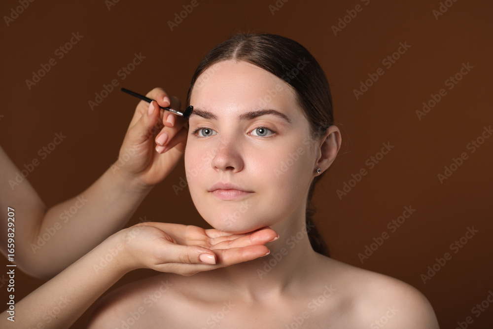 Fototapeta premium Cosmetologist brushing young woman's eyebrow with spoolie on brown background, closeup