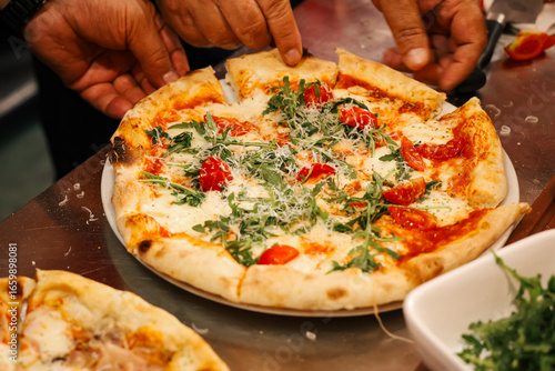 Chef’s hands preparing and serving freshly baked pizza with arugula and cherry tomatoes in a busy kitchen, authentic culinary work atmosphere