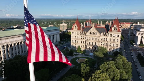 Waving American flag on flagpole at west capitol park in Albany. Aerial closeup. Chancellor hall and New York State capitol building in distance. Golden hour in the evening. Summer season.
