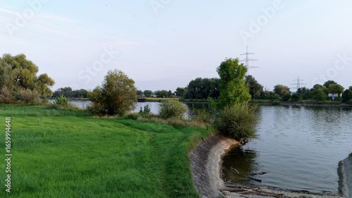 Drone flight at low altitude over a green meadow on the banks of a wide river at sunset. The light highlights the natural structure of the landscape and creates a calm and peaceful view of the scenery