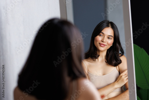 Happy beautiful young lady touching face look in bathroom mirror