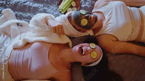 Cheerful female friends wearing bathrobes relaxing together, applying face masks and placing cucumber slices over eyes during home-based wellness and beauty pampering session
