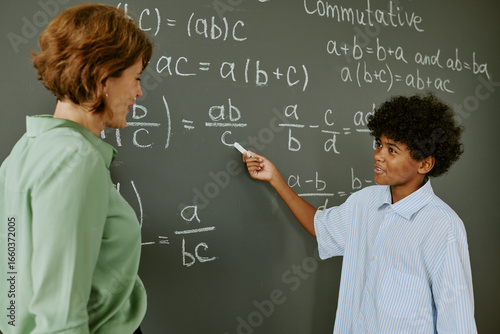 Quadro su tela Black teenage boy explaining math equation on chalkboard while middle aged Cauca