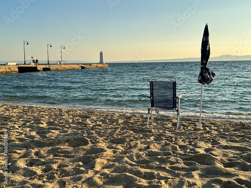 A tranquil sandy beach at sunset with a single chair and umbrella facing the sea, near a pier with lanterns and a distant lighthouse