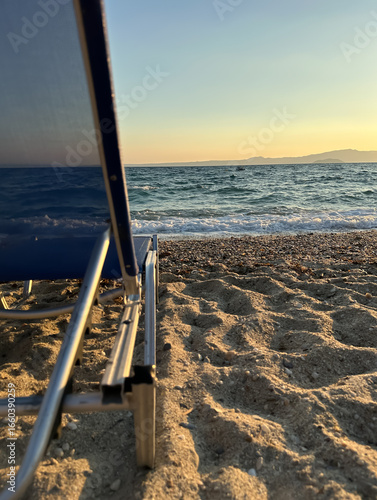 A beach chair on sandy shore faces the calm sea at sunset, capturing a tranquil summer moment