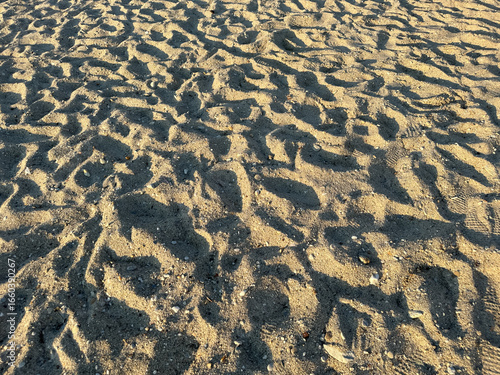 Golden sunlight casts deep shadows across textured sand covered with footprints on the beach