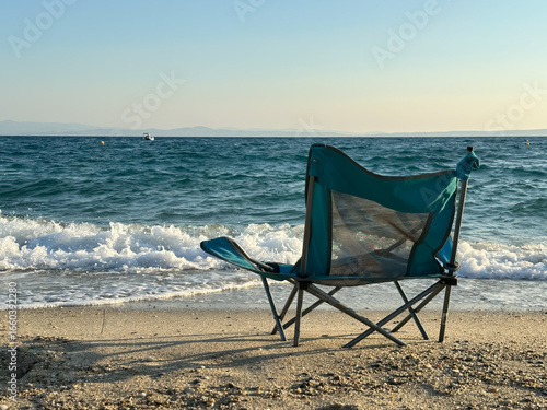 A folding beach chair stands on the sandy shore facing the turquoise sea waves under the afternoon sun