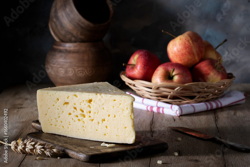 Piece of cheese on cutting board and apples in wicker plate on rustic table