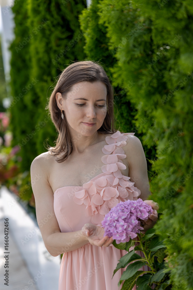 Fototapeta premium Young woman in an elegant light pink dress with floral details gently holding a blooming purple hydrangea, standing beside lush green bushes in a summer garden
