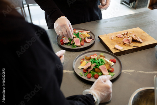 In the restaurant kitchen, the cooks are preparing a warm salad with roast beef. On the plates are fresh vegetables, pieces of meat and cheese. Sliced ​​ingredients are lying on the board nearby. 