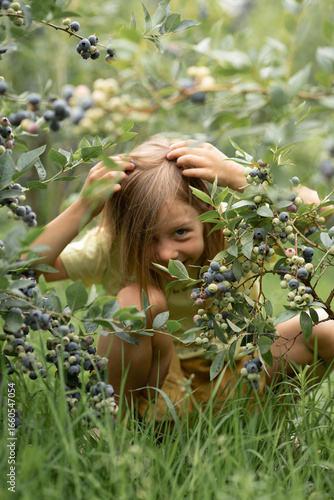 a little girl is hiding in a bush with blueberries, and she has a blueberry on her head