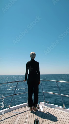 Elegant aerial drone video shows a gender-neutral, diverse individual at a sleek modern yacht's bow, gazing serenely at the shimmering ocean under a blue sky with warm sunlight. Aspirational luxury,