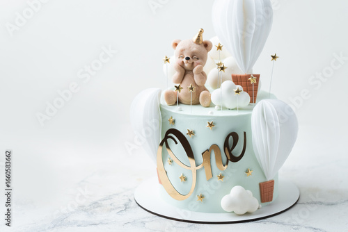 Birthday cake frosted with turquoise chocolate velvet coating and decorated with edible teddy bear figure and balloons on top. Cute birthday cake for a little one year old baby boy on white background