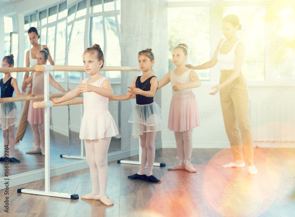 Fototapeta premium Group of ballerinas preparing for performance, learning and improving quality of rendering stage. Ballerinas near ballet bare repeat first position with supervision experienced female mentor