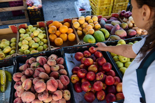 A woman selects vibrant fresh fruits at an outdoor market stall. The array of colorful produce, including peaches, apples, and bananas, highlights healthy food choices and local produce availability.