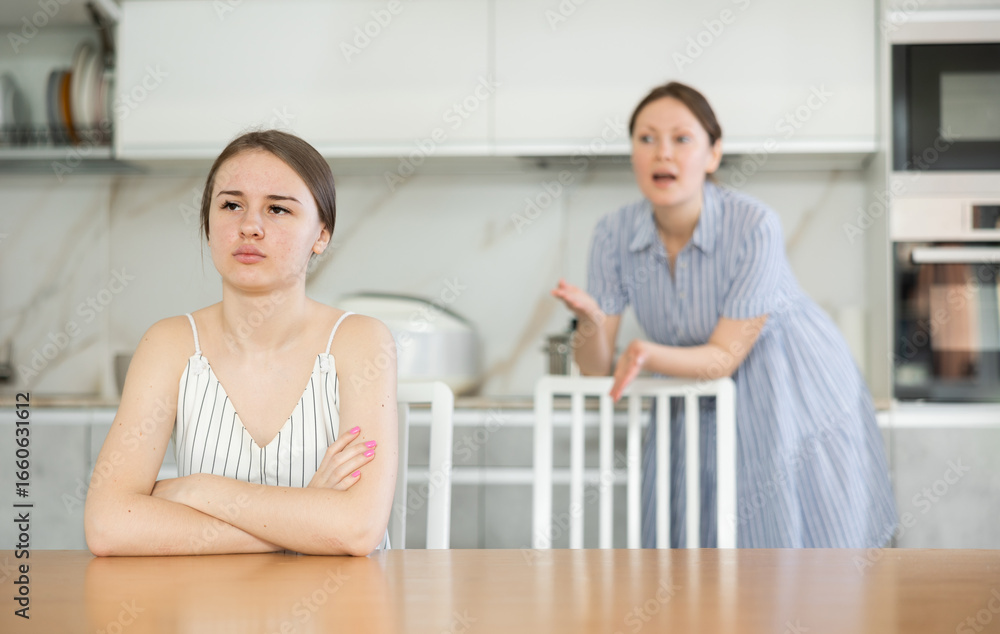 Fototapeta premium Upset teenage girl sitting at the kitchen-table turning her back upon mother attempting to come to terms with daughter