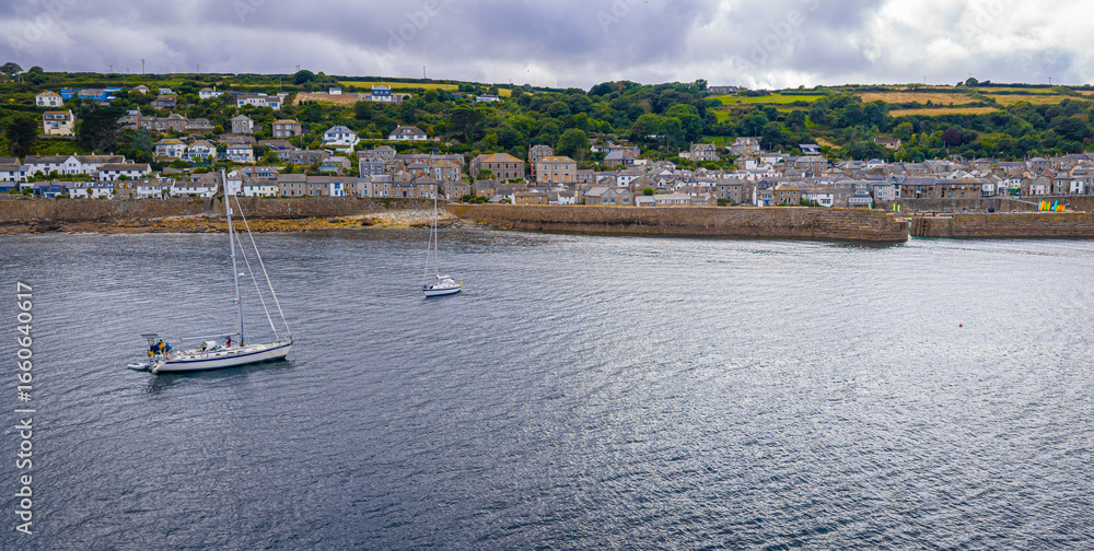 Obraz premium Aerial view of Mousehole harbour in Cornwall, UK, showing moored boats, turquoise waters, sandy shore, and surrounding stone buildings