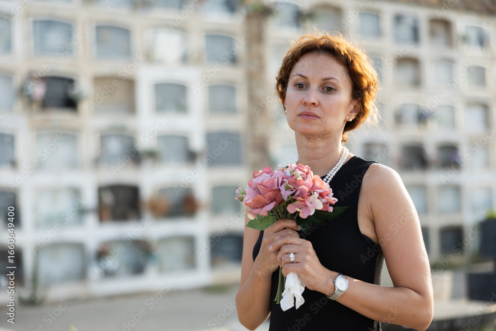 Fototapeta premium Sorrowful young woman in black dress with bouquet of flowers and handkerchief in hands, stands in cemetery. Visiting grave of deceased relative, mourning dead loved one. Appeal to God, prayer for dead