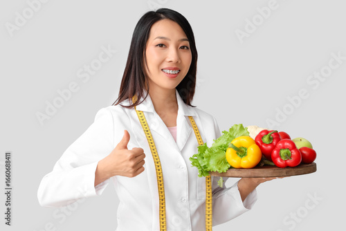 Female Asian nutritionist with healthy food showing thumb-up on light background