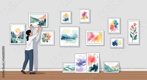Woman hangs various framed artworks on a light gray wall, showcasing a collection of abstract and landscape paintings.