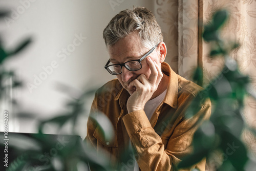 man sits at desk in his home, deep in thought with his hand on his chin. green plants frame scene, creating serene atmosphere for his focused work. close up.