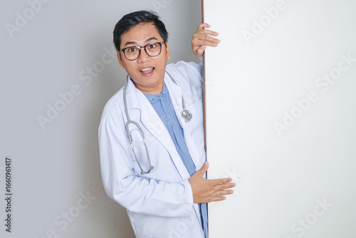 Smiling young male doctor gesturing with one hand while playfully peeking from behind blank wall, ideal for interactive and engaging healthcare advertising