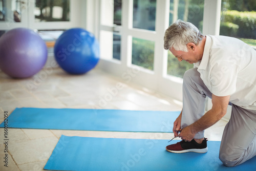 Man kneeling tying sneaker laces on mats in home gym with stability balls near windows
