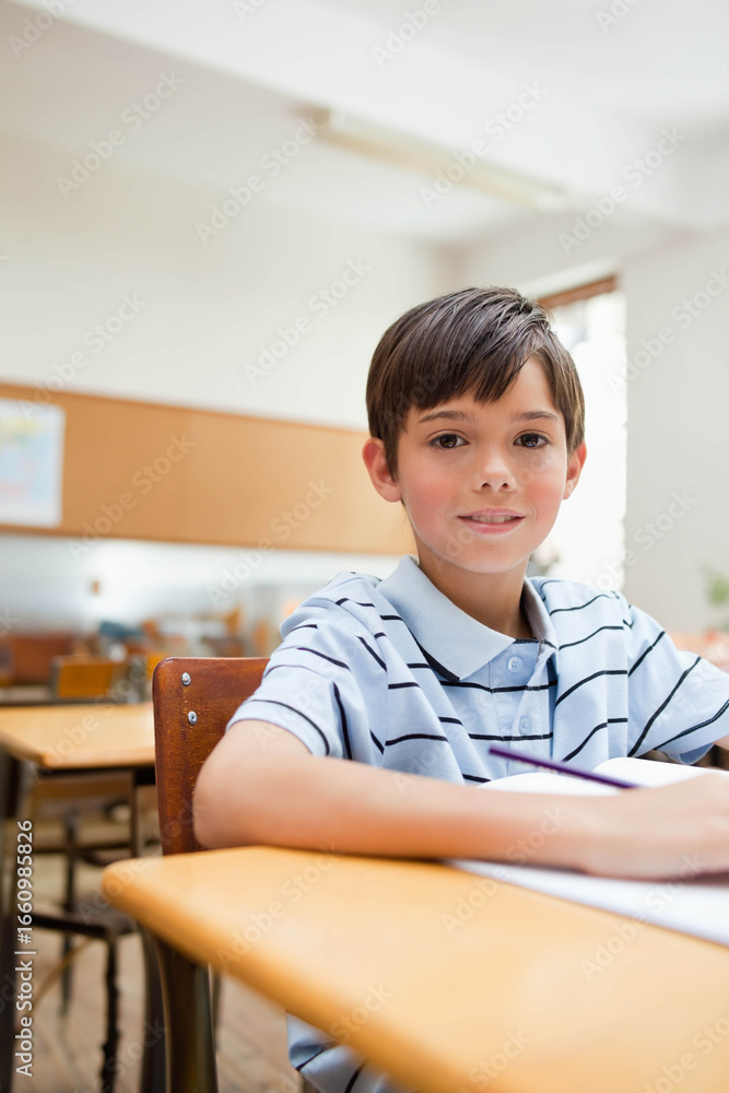 Fototapeta premium White boy student sitting at wooden desk in classroom, writing in open notebook with pencil