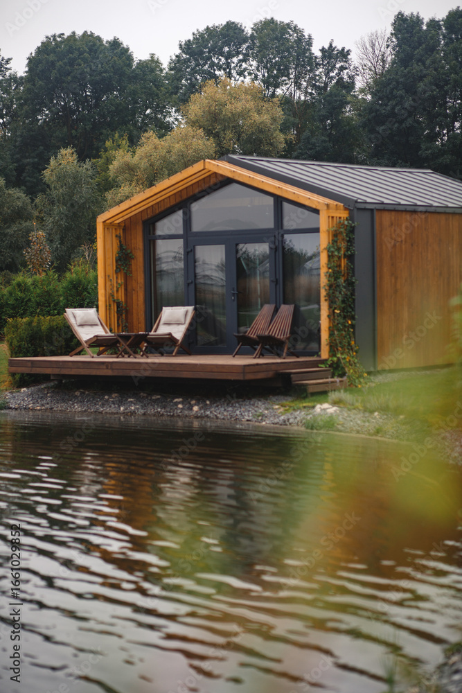 Obraz premium A modern wooden cabin with a glass facade and an outdoor deck with chairs, reflected in a calm lake with blurred plants in the foreground.