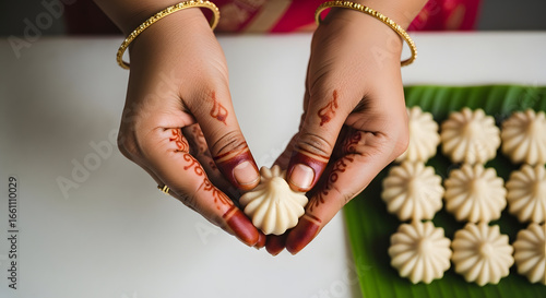 Hands decorated with henna holding an Indian sweet called modak on a green leaf with more modaks in the background