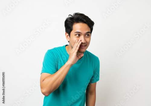 A young man in a teal shirt whispering with his hand near his mouth on a white background shot