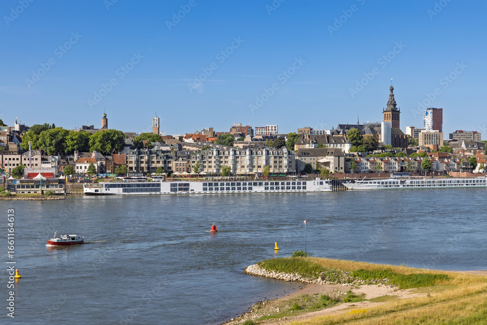 Obraz premium A picturesque panorama of Nijmegen's historic waterfront along the Waal River, with boats and city skyline.
