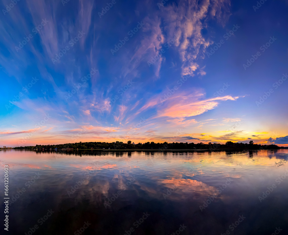 Fototapeta premium Sunset reflected in the river. Orange and pink fan clouds scatter. Calm evening river reflects the sky. Wide panoramic view of water and vegetation. Fresh water reserves. Water quality for bathing.