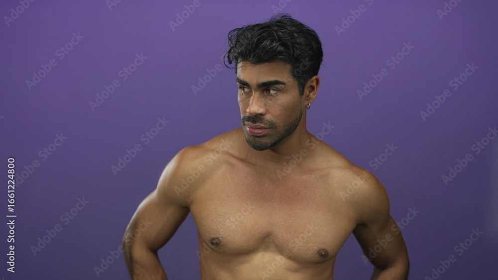 Man bare muscular chest looking down with hands on hips in a studio against a purple wall; contemplation. Man bare muscular chest looking down with hands on hips in a studio against a purple wall; contemplation.