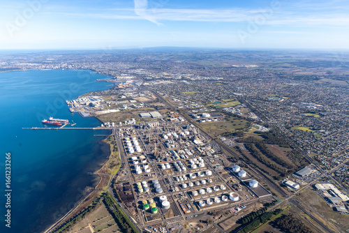 Aerial view of industrial tanks gleaming under the sun near the shimmering Corio Bay, contrasting with the lush green suburbs stretching towards the horizon, Geelong, Victoria, Australia.
